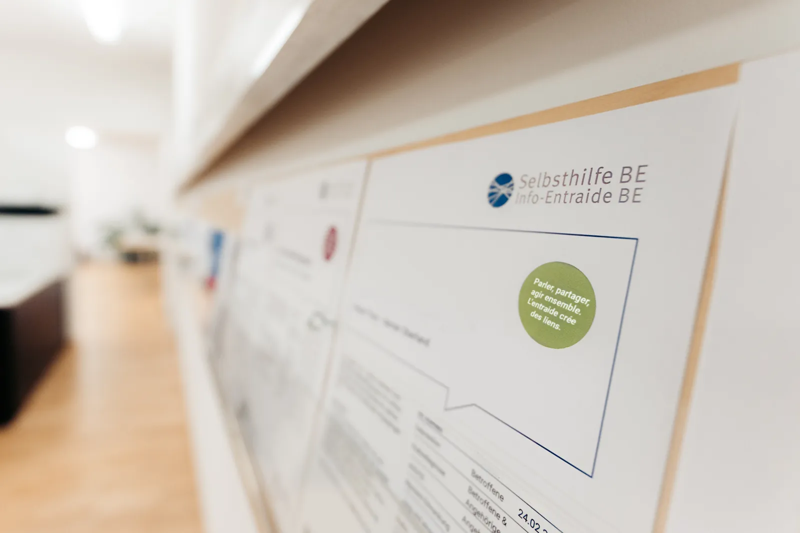 Un gros plan sur un tableau d'affichage dans un couloir lumineux. Au premier plan, un flyer de l'organisation « Selbsthilfe BE / Info-Entraide BE » avec un autocollant vert rond portant l'inscription en français : « Parler, partager, agir ensemble – L’entraide crée des liens ». L'arrière-plan est flou et montre d'autres documents ainsi qu'un sol en bois.