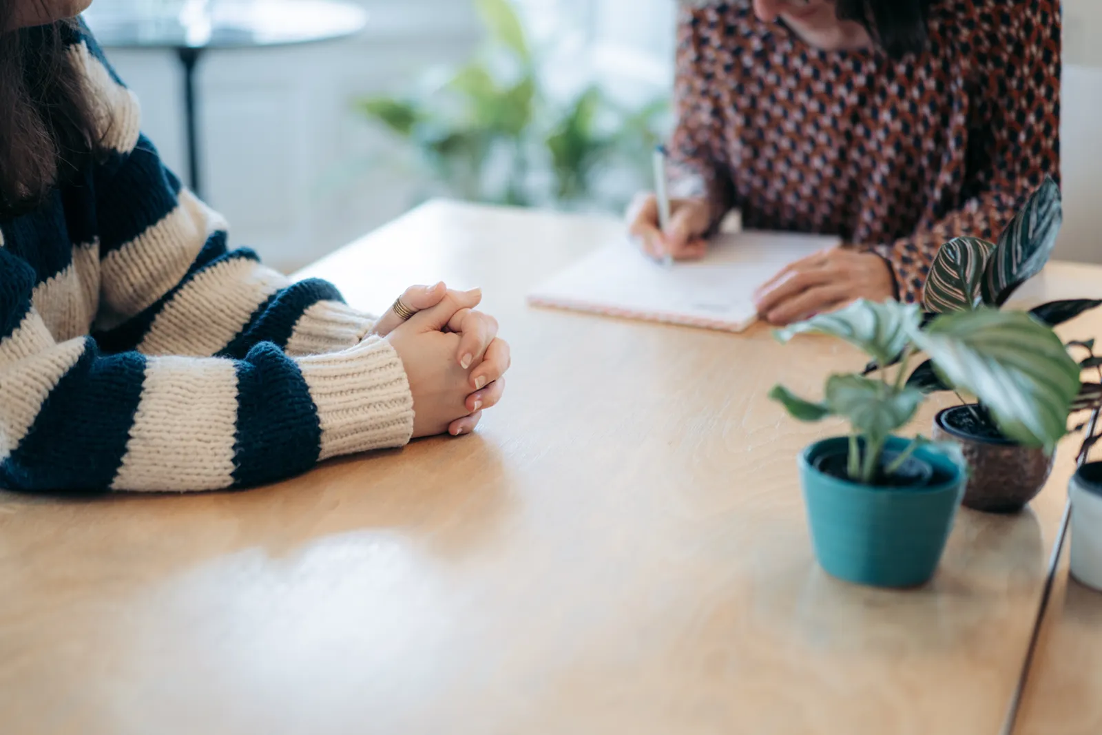 Une scène de consultation : Deux personnes sont assises à une table en bois. La personne au premier plan, portant un pull à rayures bleues et blanches, écoute avec les mains croisées. En arrière-plan, la conseillère prend des notes dans un carnet. De petites plantes sur la table créent une ambiance détendue.