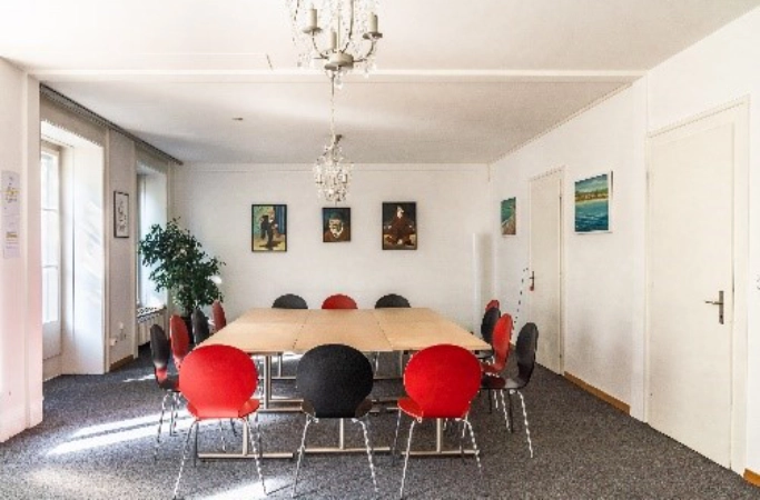 Salle de groupe avec des tables rectangulaires en forme de U, des chaises rouges et noires, des tableaux muraux, une moquette et des lustres.