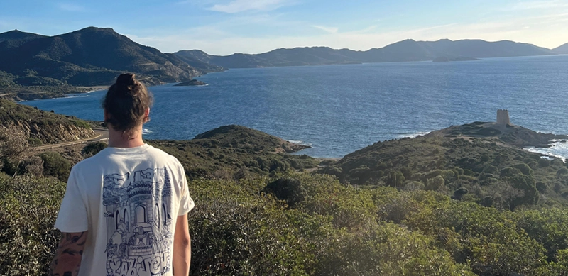 ersonne avec chignon et t-shirt à grand motif vue de dos regardant un paysage côtier avec collines, mer et une tour sur une presqu'île