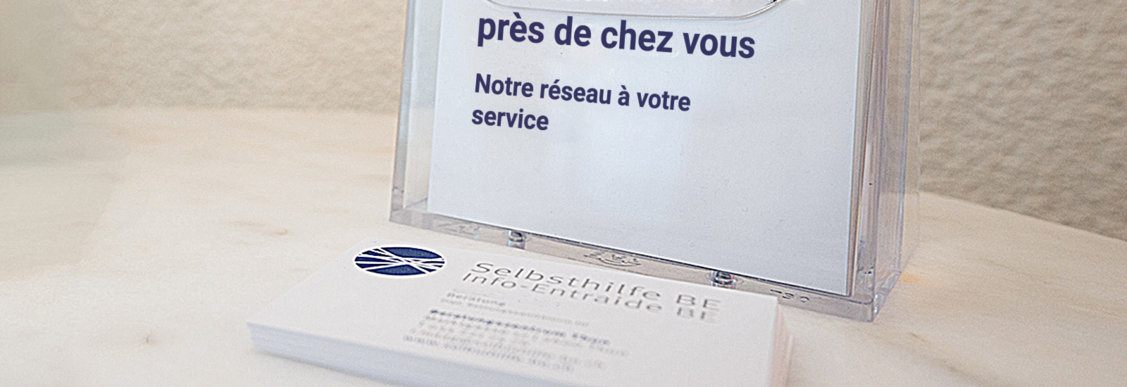 Une pile de cartes de visite de l'organisation « Info-Entraide BE » posée sur une table en bois clair. Derrière se trouve un présentoir en acrylique transparent avec un insert portant l'inscription en grand : « près de chez vous » et en dessous « Notre réseau à votre service ».