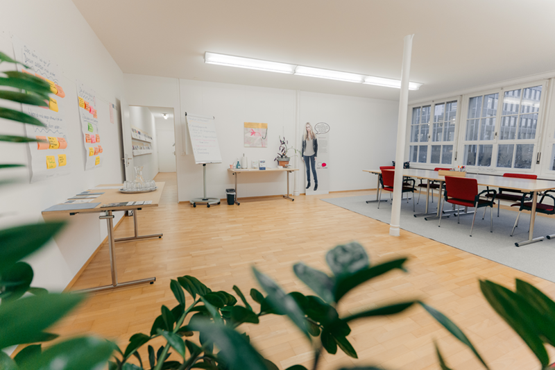 Salle de séminaire moderne avec parquet, grandes fenêtres, table rectangulaire, chaises de différentes couleurs et un flipchart.