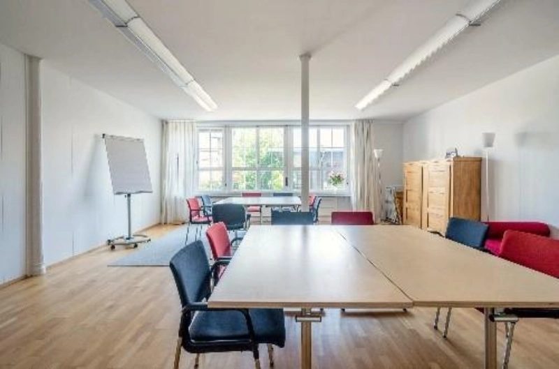 Salle de séminaire moderne avec parquet, grandes fenêtres, table rectangulaire, chaises de différentes couleurs et un flipchart.
