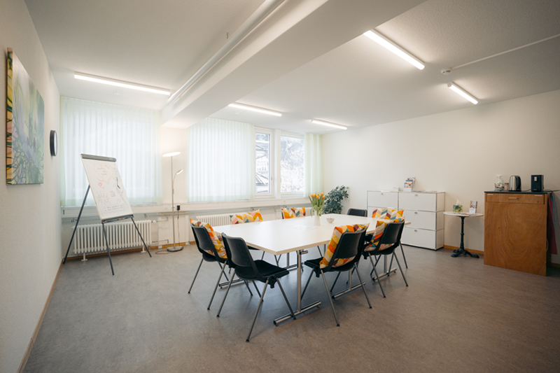 Salle de groupe lumineuse avec une grande table rectangulaire, six chaises noires, des coussins colorés sur les rebords de fenêtre et des plantes d’intérieur.