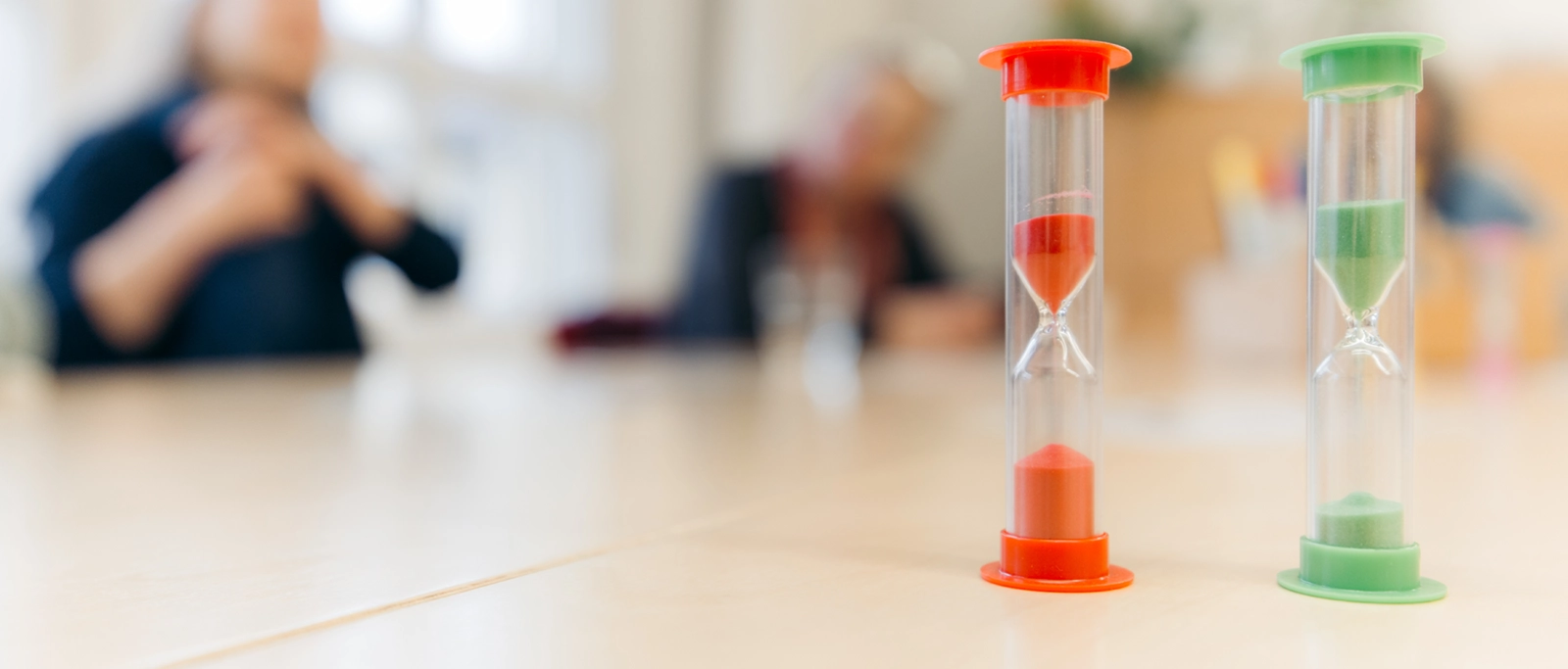 Deux sabliers avec du sable rouge et vert posés sur une table devant des personnes floues en arrière-plan.